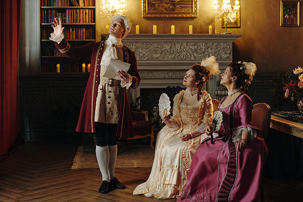 Caucasian middle aged man in Baroque costume gesturing dramatically while holding paper, two Caucasian middle aged women in elaborate Renaissance dresses sitting and watching him