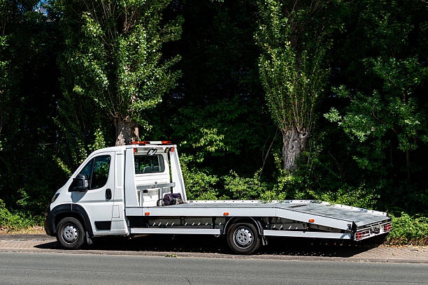 A white flatbed tow truck is parked along a quiet road adjacent to a lush area filled with green trees under clear skies. The truck appears ready for transport or hauling tasks.