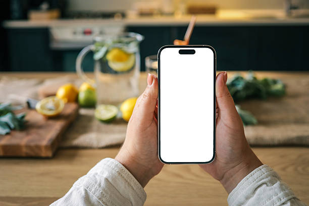 istock Hands holding a smartphone with blank white screen in kitchen, lemon water and herbs blurred in background. Mockup for app, website, or social media content 2226521499