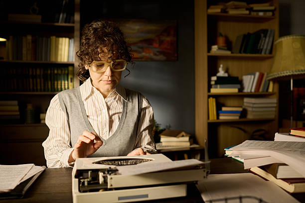 Middle aged Caucasian woman typing on vintage typewriter in office surrounded by stacks of documents and books, wearing glasses and focusing on work at wooden desk