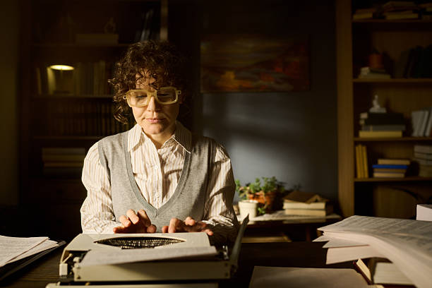 Caucasian middle aged woman typing on typewriter in office surrounded by stacks of papers and books, wearing glasses and focusing on work, bookshelf and painting in background