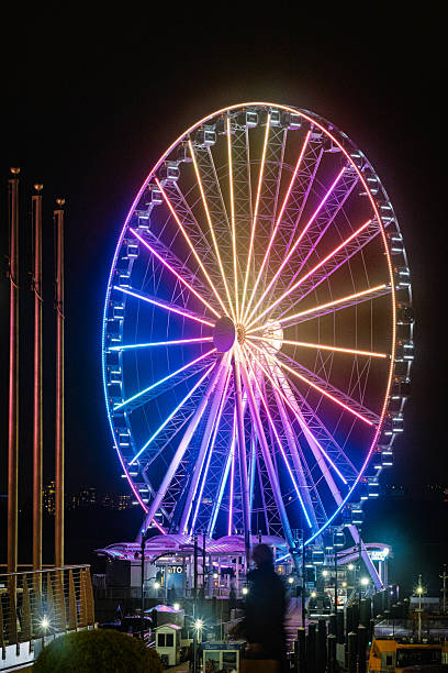 This striking vertical shot captures the Capital Wheel at National Harbor glowing with vibrant, multi-colored lights against a dark night sky. The Ferris wheel's spokes and cabins are illuminated in shades of blue, pink, yellow, and orange, creating a spectacular light display. In the foreground to the left, tall golden poles rise, and below, the bustling activity of the harbor is suggested by lights and subtle details of the surrounding structures. The image is a dazzling portrayal of a popular amusement attraction after dark. High quality photo