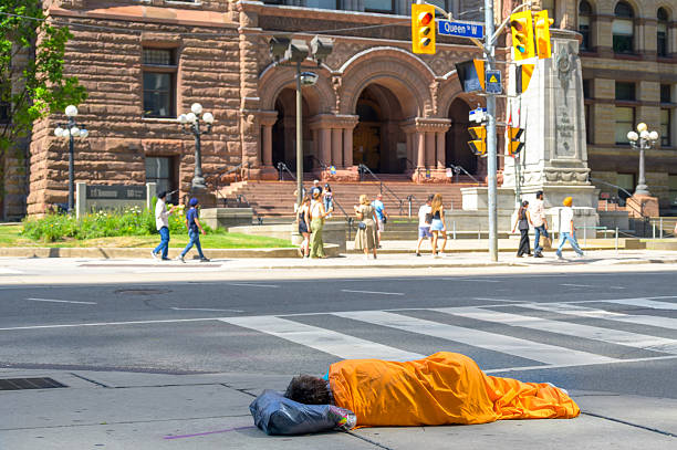 Toronto, Canada - July 13, 2025: Homelessness as a social issue. A person sleeps on the sidewalk wrapped in an orange blanket near a crosswalk and heritage building.