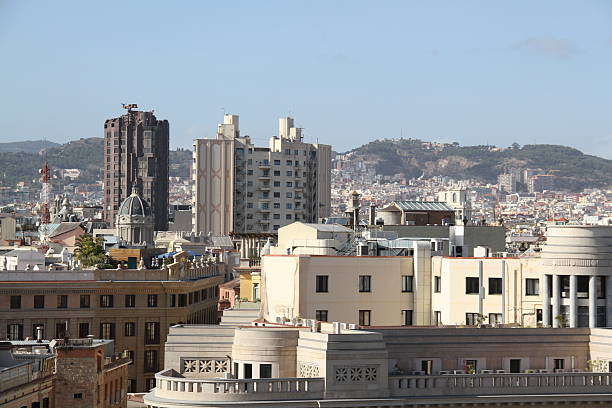 A sweeping rooftop view of the Barcelona skyline on a bright summer day reveals a vibrant cityscape dotted with terracotta rooftops, historic spires, and modern high-rises. The Mediterranean light casts crisp shadows, while the distant blue of the sea and sky frames the urban expanse in a warm, sunlit glow.