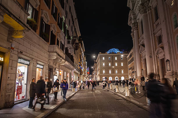 Rome, Italy - January 15, 2025: Night photograph of Via del Corso, one of Rome's best known shopping corridors through the historic center. The camera looks along the broad street lined with illuminated retail facades including fashion stores, seasonal sale signage and flags. Shoppers carrying bags move in both directions, some blurred by motion, conveying evening activity. At the far end historic palazzi and church architecture frame the view, with a modern glass dome lit in blue above the rooftops. The scene documents Rome nightlife, consumer activity and pedestrian flow on a major high street.