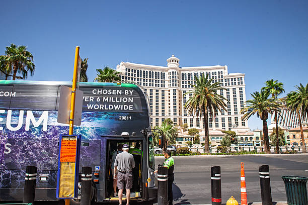 Las Vegas, August 21, 2024: The moment of boarding is frozen in this street level view of The Deuce, the air conditioned double-decker bus that shuttles visitors up and down the Las Vegas Strip twenty four hours a day. Seen against the fountain lined facade of the Bellagio and a row of mature palms, a grey capped rider climbs the rear stairs while a uniformed transit security officer watches the platform, highlighting safety procedures. The exterior wrap promoting Arte Museum adds swirling blues and purples that pop under midday sun, while reflective windows mirror the roadway. Bright detour signage, bollards fitted with mobility symbols and a lowered kneeling feature demonstrate accessibility for elderly and disabled passengers. The photograph blends human interest and urban detail, offering material for stories about public transportation, sustainable tourism, fare systems or the daily life of service staff who keep the Nevada economy moving.