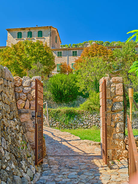 A picturesque courtyard featuring antique stone architecture surrounded by vibrant greenery, with an open gate leading into a traditional rustic house in Majorca. The setting radiates tranquility and Mediterranean charm.