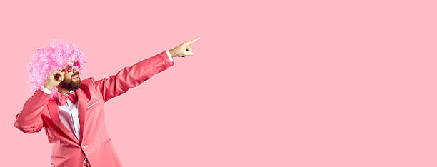 Portrait of cheerful guy points at blank text copy space background on the right. Funny clown wearing funky pink suit, curly carnival wig and sunglasses invites you to cool party or great holiday sale