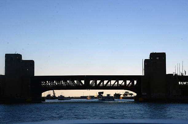 A silhouetted of the Lake Shore Drive bridge stretches across a body of water at sunrise in Chicago, Illinois, its truss design and vertical supports casting bold shadows against a gradient sky. Boats drift beneath the span, while distant towers frame the scene, evoking themes of architectural contrast, maritime rhythm, and civic transition. in Chicago, IL, USA on May 25,2025