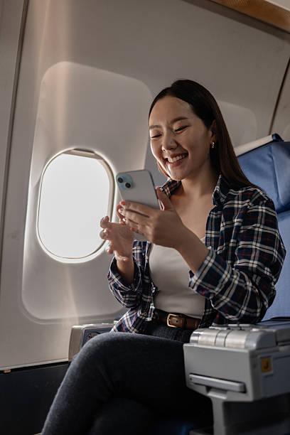 Asian woman smiling while using her smartphone on an airplane. She is sitting next to the window, enjoying her time during the flight. The atmosphere appears relaxed and comfortable.