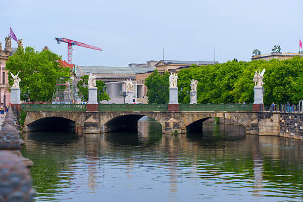 Berlin, Germany - June 1, 2025: Scenic view of a historic bridge adorned with statues, spanning a calm river in Berlin, surrounded by lush green trees and architectural landmarks.