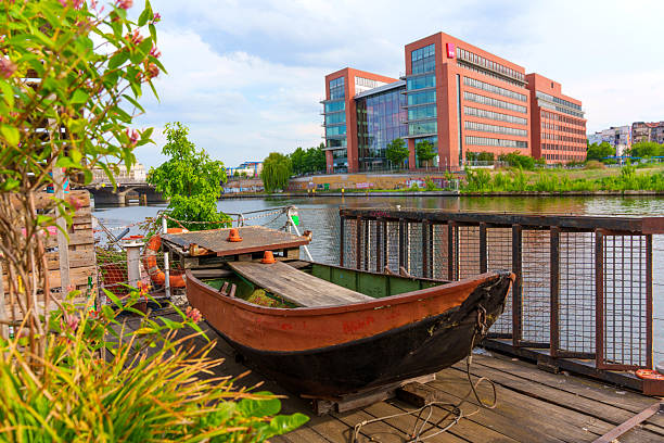 A weathered iron mooring ring is mounted on the riverside promenade along the Spree River in Berlin, Germany. In the background, a modern yacht glides past urban buildings and green trees under a clear blue spring sky. The photo captures a contrast between industrial infrastructure and leisurely boating.