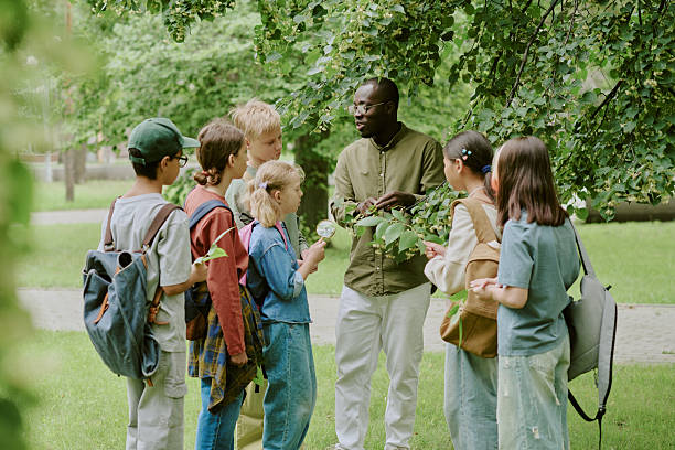 A diverse group of children and a supervising adult walk along a sunny meadow, sketching, writing, and sharing notes as they explore nature together.
