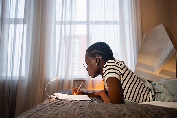 istock Young black woman lying on bed writing in notebook by lamp light 2226600105