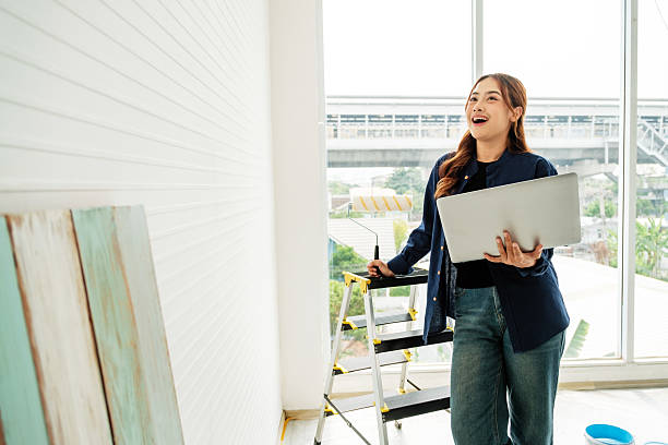 Young woman plans a home improvement project while holding a laptop and paint roller, standing by a bright window. The scene captures creativity and modern DIY renovation in natural light.