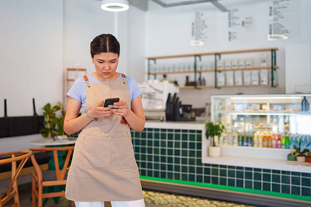 A young female cafe employee is seen using her mobile phone. The scene captures her engagement with technology, possibly for communication, checking orders, or managing cafe operations in a bustling environment.