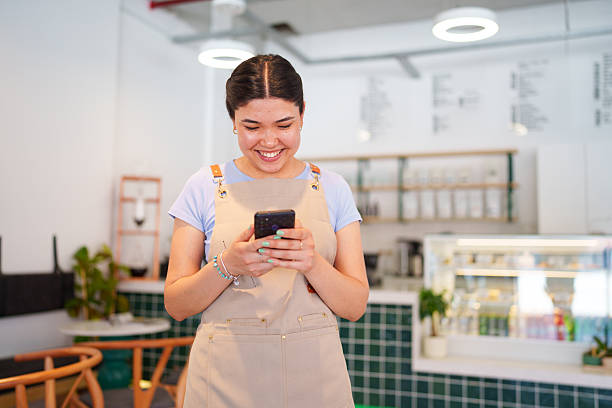 A young female cafe employee is seen using her mobile phone. The scene captures her engagement with technology, possibly for communication, checking orders, or managing cafe operations in a bustling environment.