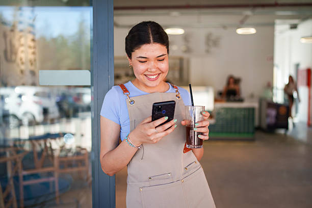 A young female cafe employee is seen using her mobile phone. The scene captures her engagement with technology, possibly for communication, checking orders, or managing cafe operations in a bustling environment.