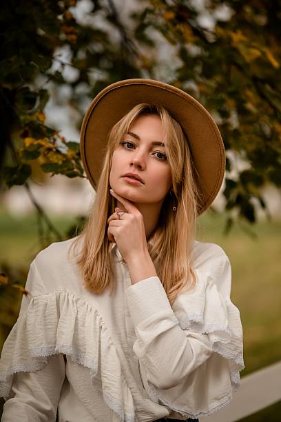 Woman in rustic setting wearing a stylish hat and ruffled blouse. Her thoughtful expression and soft autumn backdrop create a sense of introspection and seasonal charm.