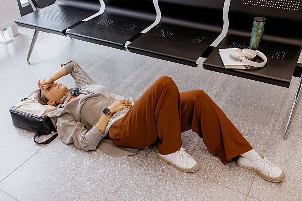 Tired woman lying on the floor in a public waiting area, resting on her backpack, feeling exhausted during long travel delay, headphones and notebook nearby
