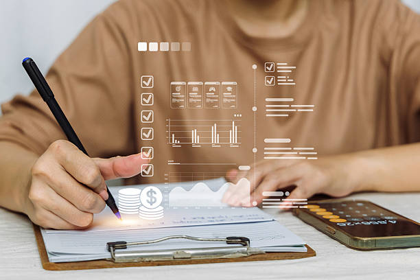 A person writes on a clipboard while viewing a complex digital dashboard for financial management and tracking.
