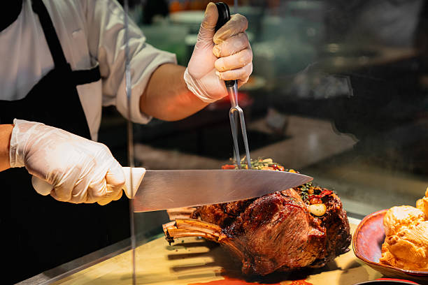 A chef wearing gloves and a black apron slices a juicy prime rib roast using a carving fork and a large knife. The scene takes place at a buffet or carving station, showcasing professional food preparation and hospitality service. Rich textures and warm lighting highlight the perfectly roasted meat.