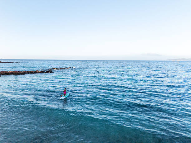 Woman in pink swimsuit paddleboarding on calm blue ocean near rocky shore. Solo water sports activity on peaceful sea. Outdoor recreation and fitness on open water.