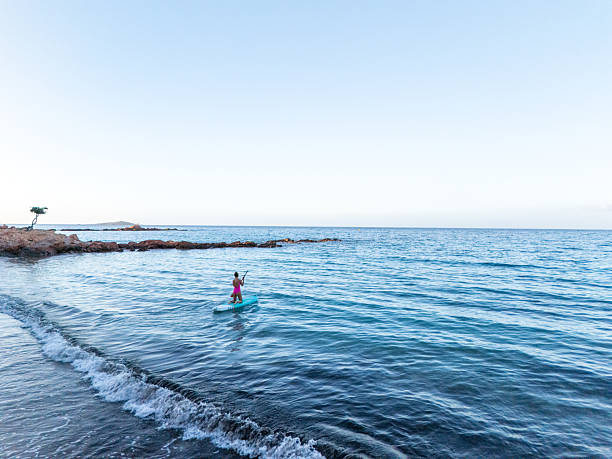 Woman in pink swimsuit paddleboarding on calm blue ocean near rocky shore. Solo water sports activity on peaceful sea. Outdoor recreation and fitness on open water.