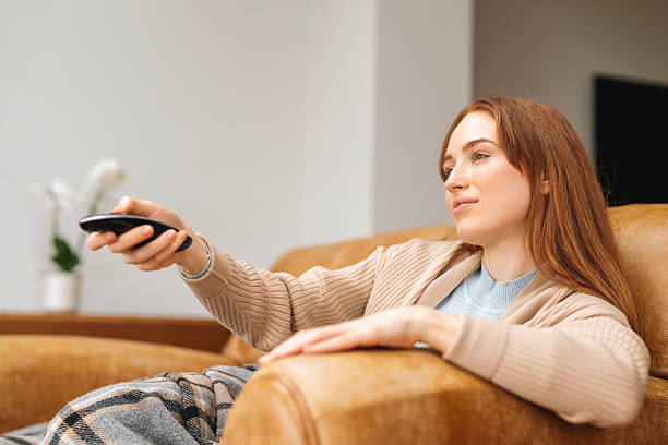 istock Woman relaxing on a sofa while watching television and using a remote control in a cozy living room decorated with indoor plants 2226635066