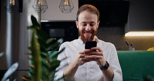 istock Happy smiling caucasian man using mobile phone resting on couch at home in kitchen. Male with three-day stubble smiles and shakes head with pleasure surprise. 2226640624