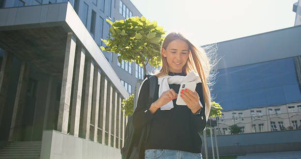 istock Good-looking satisfied smiling young light -haired girl in casual clothes with backpack standing near office building and uses her mobile on sunny day 2226645182