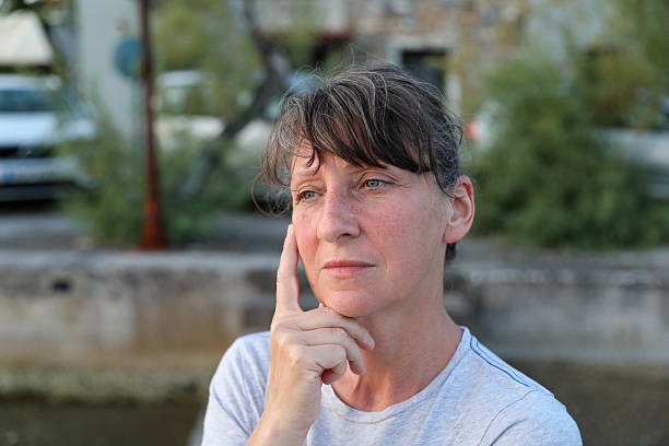 Mature Woman in Her 50s Looking Pensive with a Finger on Her Cheek, Standing Outdoors in Natural Light, Expressing Contemplation and Calmness