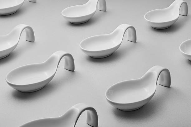 A grayscale photograph displays a pattern of identical, curved spoon-shaped ceramic dishes arranged on a flat, light gray surface. The dishes are uniformly spaced and overlap slightly