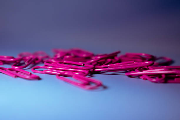 Pink paperclips scattered on a blue surface. The paperclips are metal and elongated with a smooth, shiny finish. Their vibrant color contrasts with the muted blue background, creating a lively visual. Suitable for use in organizing documents, they are common in school and office settings for binding papers together. The arrangement is casual, suggesting a ready-to-use state.