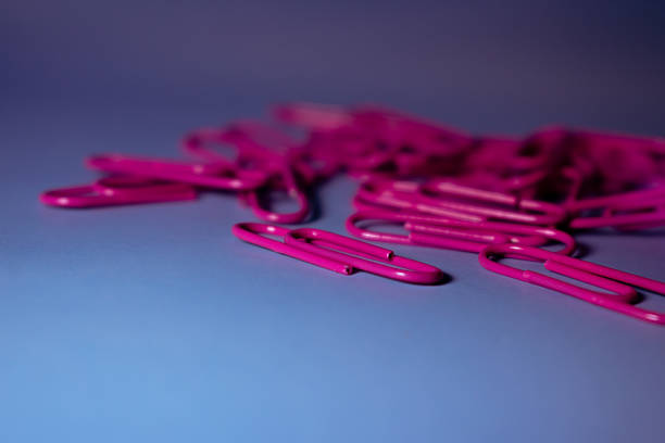 Pink paperclips scattered on a blue surface. The paperclips are metal and elongated with a smooth, shiny finish. Their vibrant color contrasts with the muted blue background, creating a lively visual. Suitable for use in organizing documents, they are common in school and office settings for binding papers together. The arrangement is casual, suggesting a ready-to-use state.