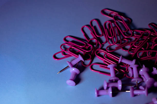 Scattered office supplies include red and pink push pins, pink paperclips, and pink binder clips on a smooth, light blue surface. The mix of colors and shapes creates a vibrant contrast against the subtle background, suitable for creative, organizational, or educational settings. The use of binder clips suggests functionality in paperwork management. The overall arrangement reflects an array of tools commonly found in school or office environments, highlighting a blend of practicality and colorful design.