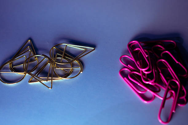 Scattered office supplies include red and pink push pins, pink paperclips, and pink binder clips on a smooth, light blue surface. The mix of colors and shapes creates a vibrant contrast against the subtle background, suitable for creative, organizational, or educational settings. The use of binder clips suggests functionality in paperwork management. The overall arrangement reflects an array of tools commonly found in school or office environments, highlighting a blend of practicality and colorful design.
