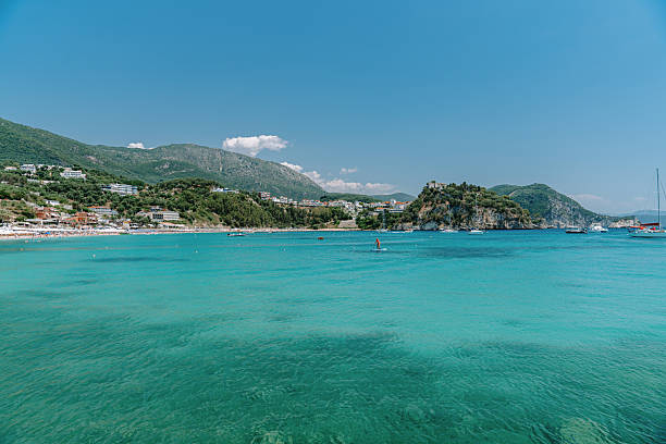 A stunning view of Valtos Beach in Parga, Greece, showcasing crystal-clear turquoise waters, lush green hills, and a vibrant coastal village. Paddleboarders and anchored boats add a touch of summer activity to this idyllic Mediterranean scene. Perfect representation of Greek island-style charm, ideal for travel, vacation, and nature themes.