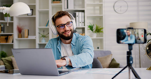 istock Handsome male tutor in headphones sitting the table writing notes by recording video class on mobile phone during online class. Concept of virtual learning, online education and people 2226663635