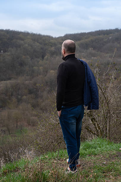 A man gazes thoughtfully over distant hills covered in trees and foliage, enjoying the tranquility of nature on a cloudy spring day while standing on a grassy slope.