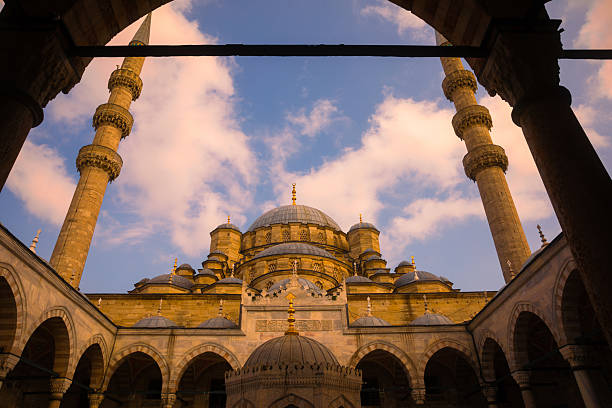 A majestic view of the Yeni Cami (New Mosque) in Istanbul, captured from within the courtyard and framed by elegant arches under a glowing sunset sky. The warm golden light enhances the details of the Ottoman architecture, domes, and twin minarets, creating a serene and timeless atmosphere. Ideal for concepts related to travel, Islamic culture, architecture, and inspirational settings.