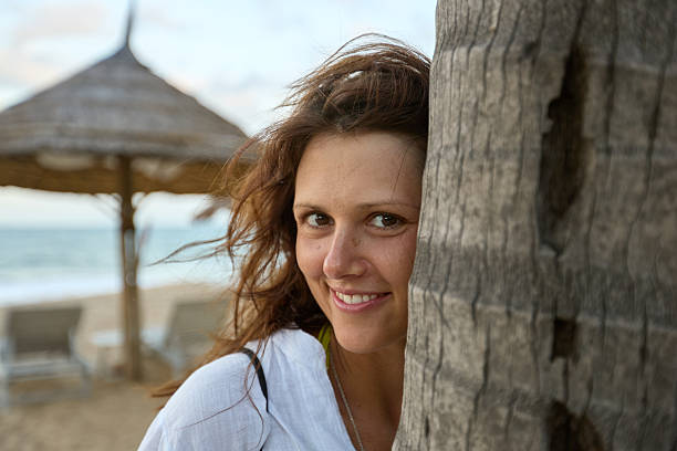 A beautiful young woman with a joyful smile peeks from behind a palm tree on a tropical beach, with a resort umbrella and ocean in the background, enjoying her vacation.