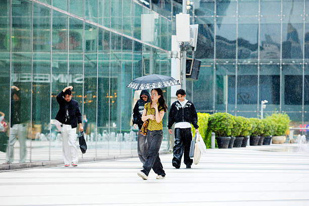 Thai woman walking with umbrella in rain on square in front of shopping mall  Siam Paragon in Bangkok downtown. Square is above sidewalk level and offers direct connection  to BTS skytrain station. Woman has eyewear and long hair. In background some young men are walking. People are on way from   Siam Center mall to BTS station and are walking along glassy facade