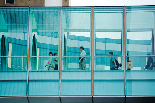 Walking people in glassy footbridge in Bangkok downtown. Footbridge is connecting the two shopping malls Siam Center and Siam Discovey.  People in scene are walking towards Siam Discovery. Some have shopping bags. Most people are wearing casual clothes. View up from sidewalk of Rama I Road