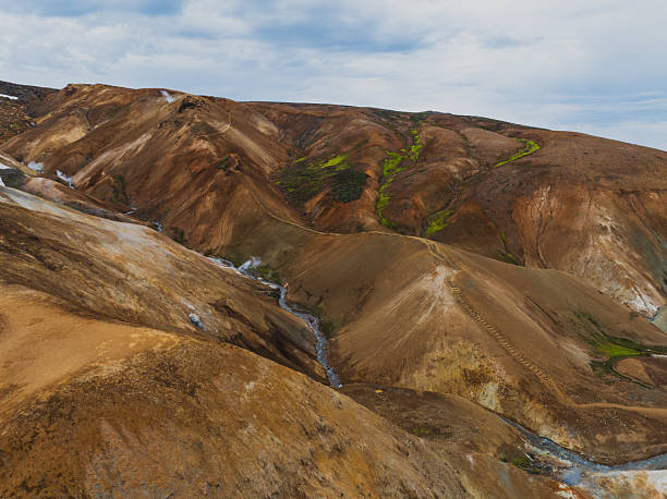 Rugged rhyolite hills in Landmannalaugar, Iceland, display warm hues, green moss, and snow patches. A small stream winds through the dramatic terrain.
