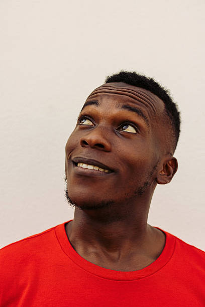 A close-up photograph of a young man in a red shirt looking upwards with a curious and joyful expression. The plain white background emphasizes the subject's face and emotion. Light creates a natural and warm atmosphere. Perfect for concepts of happiness, curiosity, contemplation, and modern casual lifestyle.