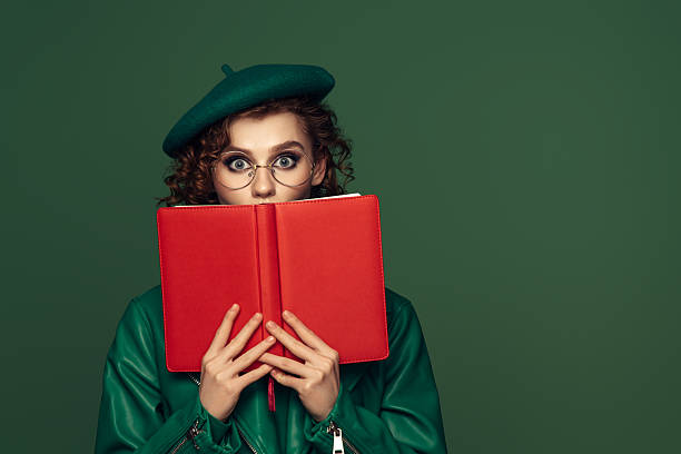 A whimsical portrait of a woman peering over a vibrant red book, dressed in a green leather jacket and matching beret. Her wide-eyed expression and round glasses add an element of intrigue and playful curiosity. The rich green background complements her outfit, while the bold red book creates a striking contrast, making the composition both dynamic and stylish.