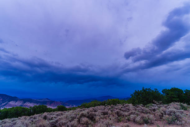 A powerful summer storm builds over the rugged mesas and shrubland of western Colorado. The dramatic sky, layered with moody purple and blue storm clouds, contrasts starkly with the arid high desert vegetation below. Captured just before dusk, the image conveys an intense, cinematic atmosphere.