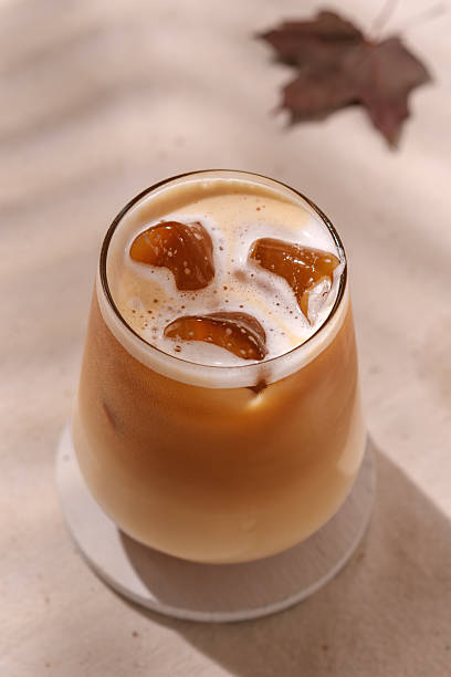 Creamy autumn latte with ice cubes glows in sunlight, maple leaf in background hints at pumpkin, apple, or maple flavors of the season.