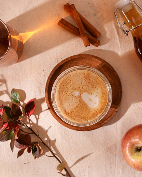 Aromatic apple latte in glass cup invites with creamy foam, surrounded by cinnamon, maple syrup, apple and autumn leaves in golden sunlight.